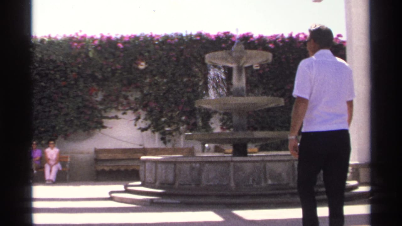 Man in a white shirt stands near a courtyard fountain while two women sit on a bench behind him.