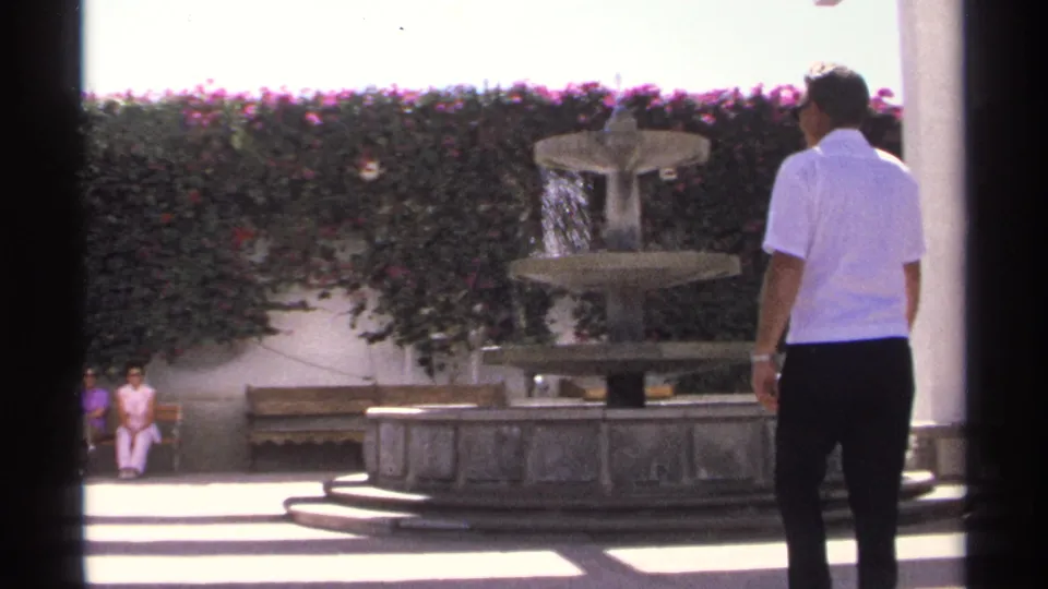 In 1975 Guaymas, Mexico, a man in a white shirt faces a tiered fountain as two women in pink sit on a bench across the courtyard.