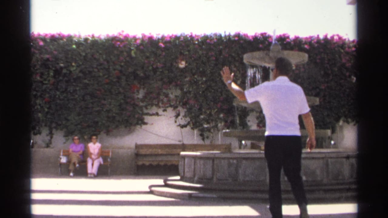 Man in a white shirt raises his hand toward two women seated near a fountain.