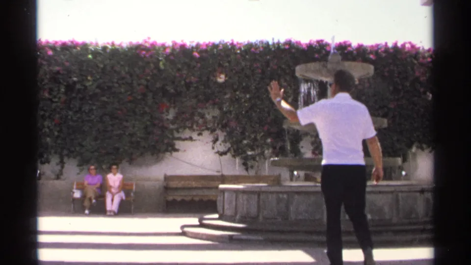 In the Guaymas courtyard, the man raises a hand in greeting toward two women sitting on a bench beside the stone fountain.