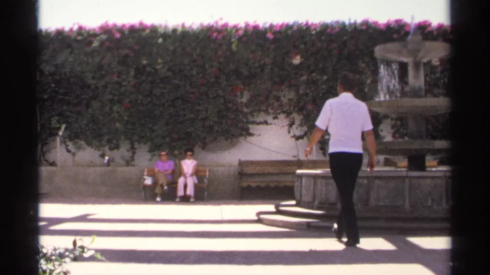 The man continues walking through the 1975 Guaymas courtyard, with the fountain at right and two women seated ahead near the hedge.