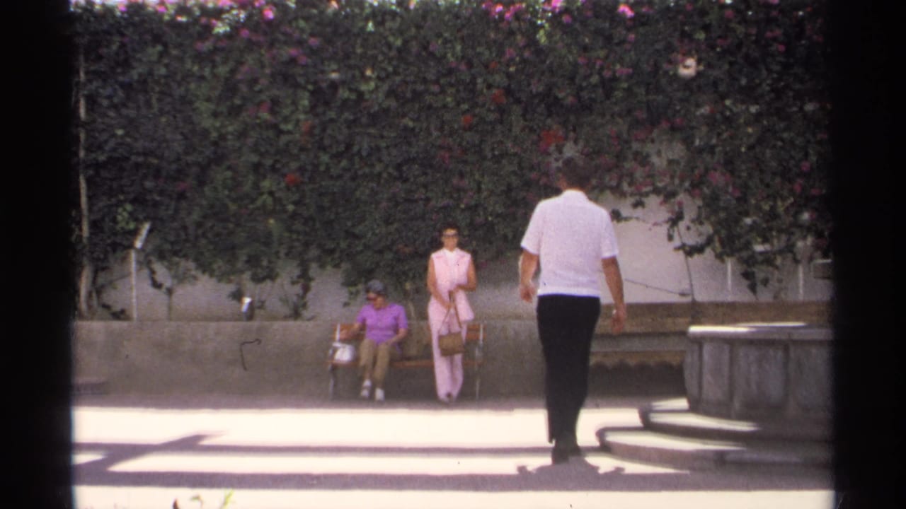 In Guaymas, Mexico, a man in a white shirt approaches two women by a wooden bench beneath a flowering hedge in 1975 8mm footage.
