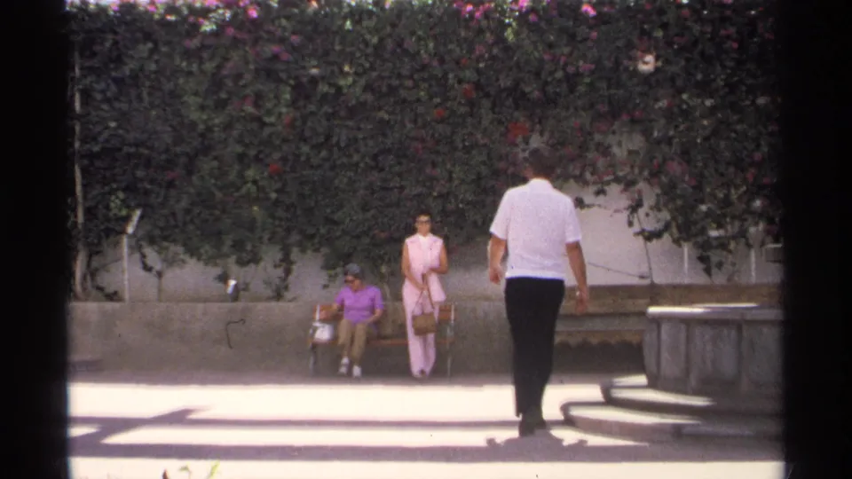 In Guaymas, Mexico, a man in a white shirt approaches two women by a wooden bench beneath a flowering hedge in 1975 8mm footage.