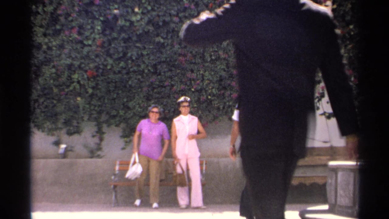 Two women stand near a bench as a suited man passes close to the camera.