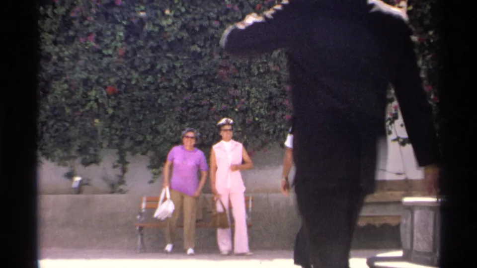 Two women in sunglasses stand near a wooden bench while a dark-suited passerby partially obscures the 1975 Guaymas home-movie view.