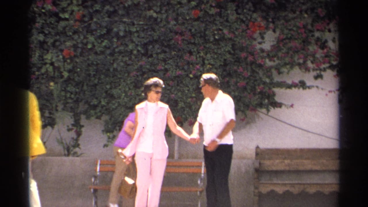 A woman in a pink sleeveless outfit stands with a man in a white shirt near a bench and flowering hedge in the Guaymas 8mm scene.