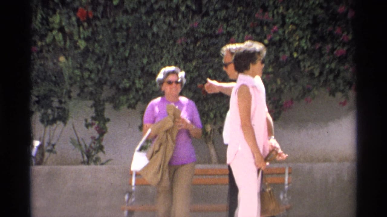 Two women, one in purple and one in pink, stand with a man near a wooden bench under a flowering hedge in 1975 Guaymas footage.