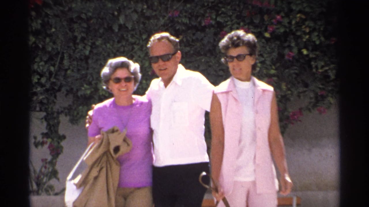 Three adults in sunglasses stand together outdoors near greenery and pink flowers.