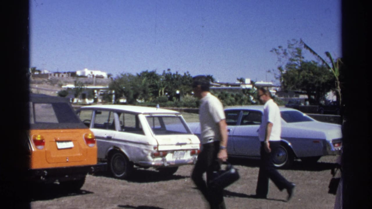 The Guaymas 1975 footage shifts to a sunny parking lot with pedestrians and several parked cars, including an orange vehicle and white station wagon.