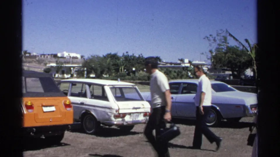 The Guaymas 1975 footage shifts to a sunny parking lot with pedestrians and several parked cars, including an orange vehicle and white station wagon.