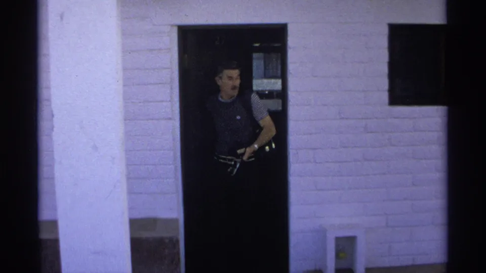 Adult man exits an apartment doorway in Guaymas, Mexico, holding a camera or small filming device in a 1975 8mm frame.