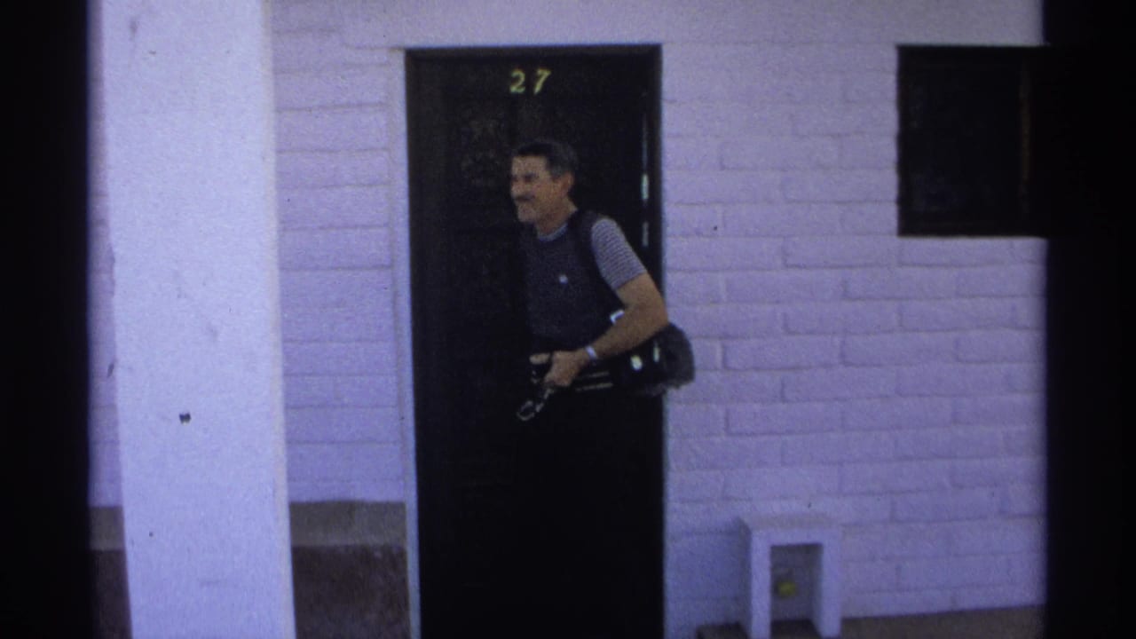 Man with camera gear stands by a dark door marked 27 beside a white brick wall.