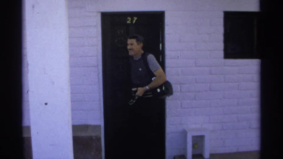 In 1975 Guaymas, Mexico, a man with camera gear stands near a numbered doorway against a white brick exterior.
