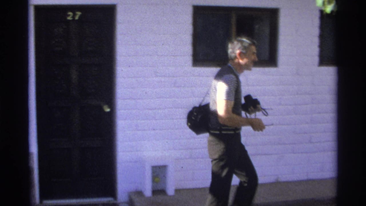 A man in a striped shirt and dark vest walks along the white building exterior with camera equipment in hand.