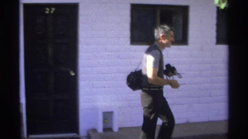 A man in a striped shirt and dark vest walks along the white building exterior with camera equipment in hand.
