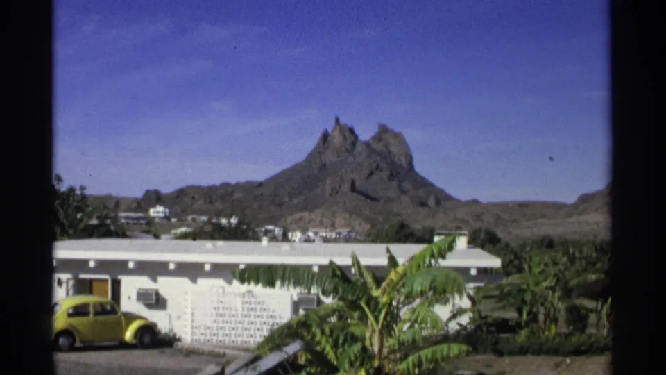 1975 Guaymas home-movie view of a rocky mountain behind low buildings, palm foliage, and a yellow car near a wall.