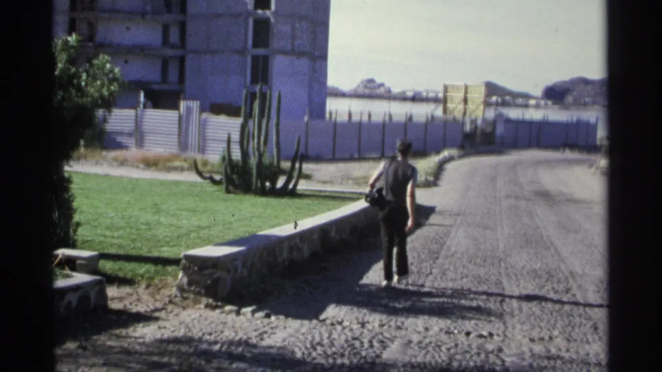 A person walks away along a rough Guaymas road near tall cactus, green grass, fencing, and an unfinished structure.