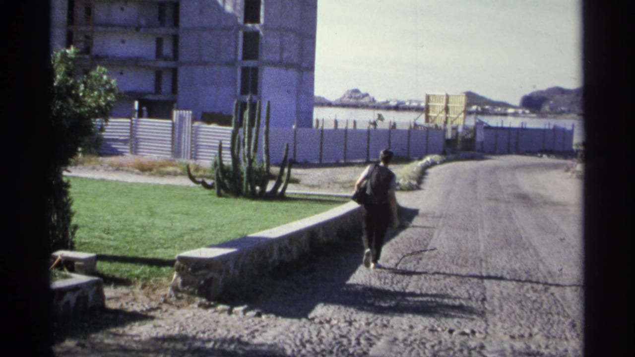 The walking figure continues away from the camera on a Guaymas road with cactus, lawn, fencing, and distant mountains visible.