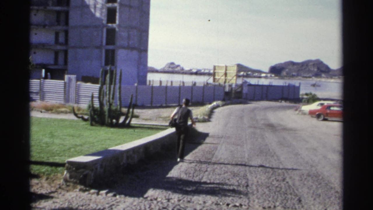 A distant person walks away on a Guaymas road framed by cactus, construction fencing, mountains, and a red car at the edge.