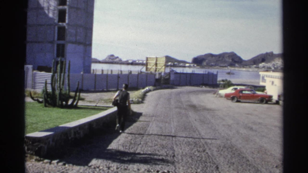 Gravel road near water with a walking adult, cactus, white buildings, and a red car.