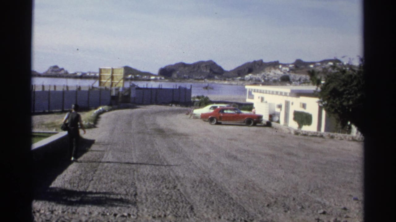 1975 Guaymas street view with a pedestrian, parked red car, low white buildings, water, and rocky hills in the distance.