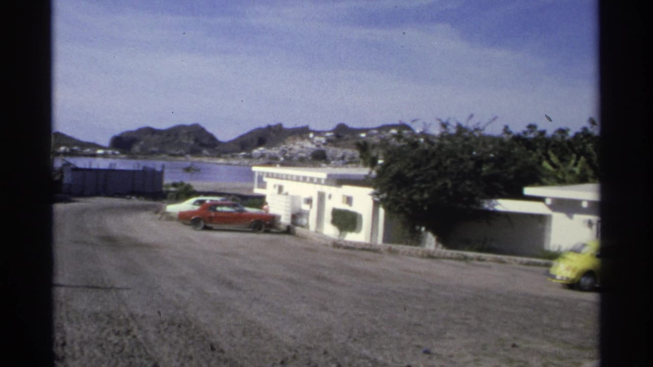 1975 Guaymas pan revealing low coastal buildings, a red parked car, distant hills, and a yellow Beetle at the edge of frame.