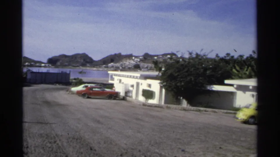 1975 Guaymas pan revealing low coastal buildings, a red parked car, distant hills, and a yellow Beetle at the edge of frame.