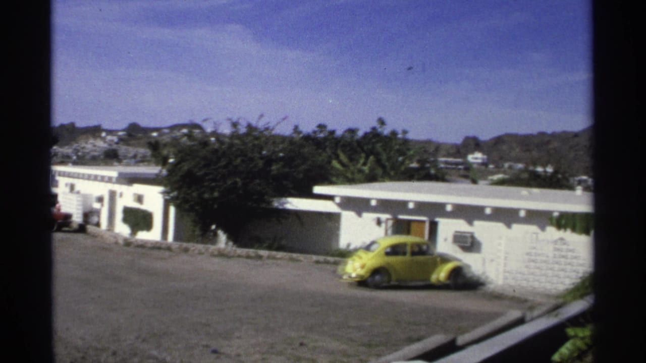 1975 Guaymas home-movie frame with a yellow Beetle near a white wall, low buildings, coastal hills, and bright sky.