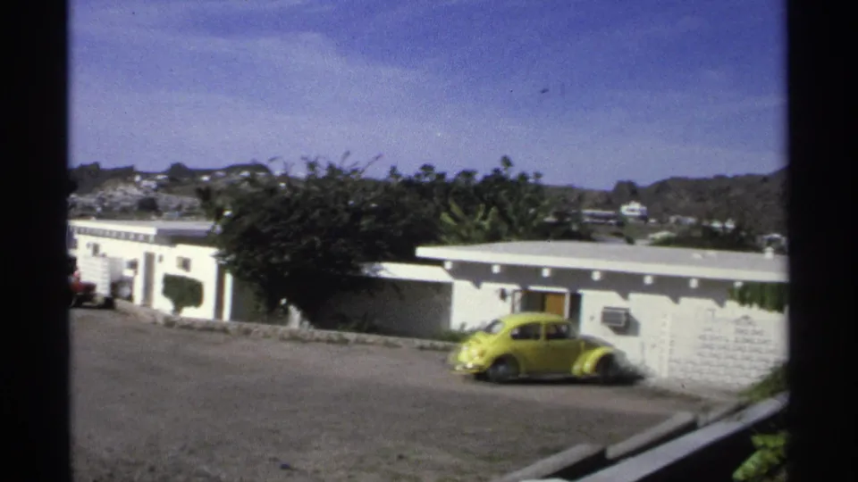 1975 Guaymas home-movie frame with a yellow Beetle near a white wall, low buildings, coastal hills, and bright sky.