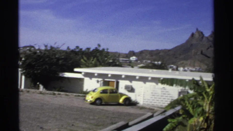 1975 Guaymas, Mexico 8mm frame showing a white low-rise residential building, yellow compact car, shrubs, palms, blue sky, and a rocky mountain backdrop.