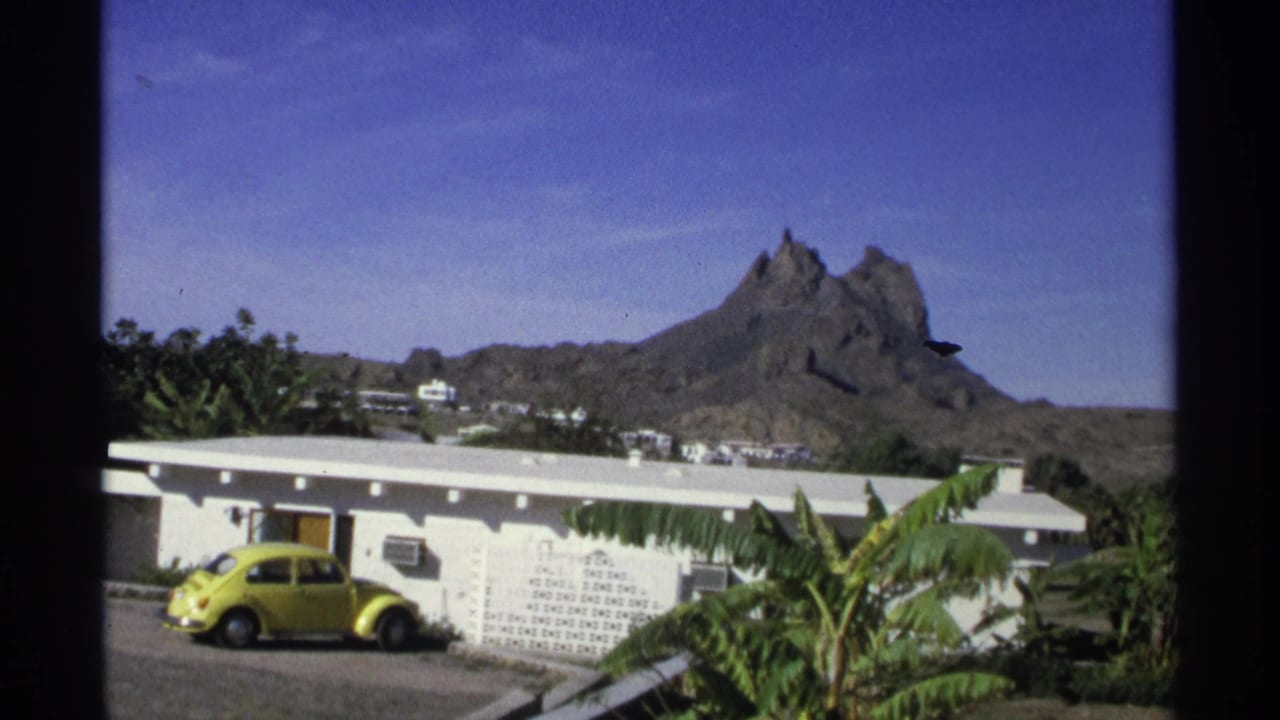1975 Guaymas, Mexico 8mm frame with a parked yellow compact car beside a white stucco building, palm plants, distant homes, and a jagged mountain ridge.