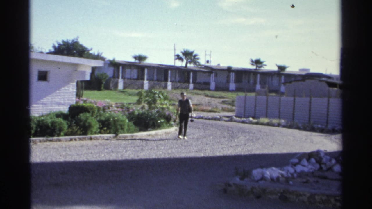 1975 Guaymas, Mexico 8mm frame showing a man walking across a gravel residential area with white low-rise buildings, shrubs, palms, and bright daylight.