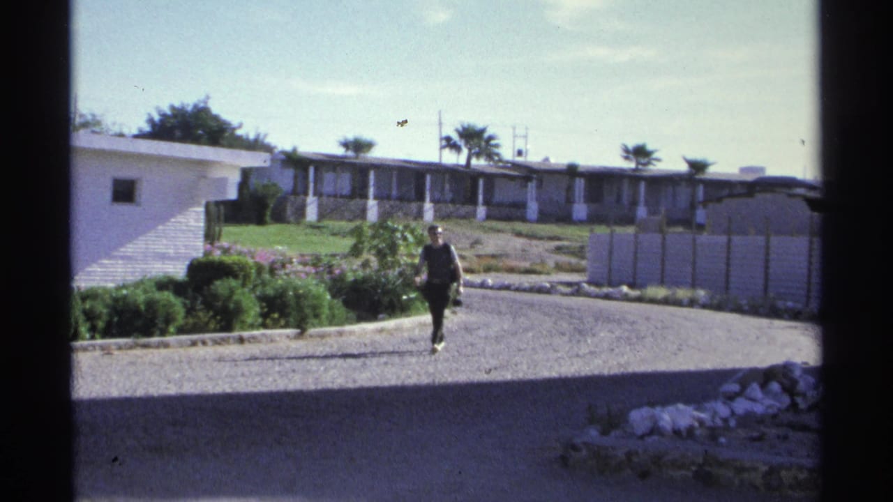 1975 Guaymas, Mexico 8mm frame of a man walking near landscaped shrubs, a gravel drive, white low-rise buildings, palm trees, and sunlit residential grounds.