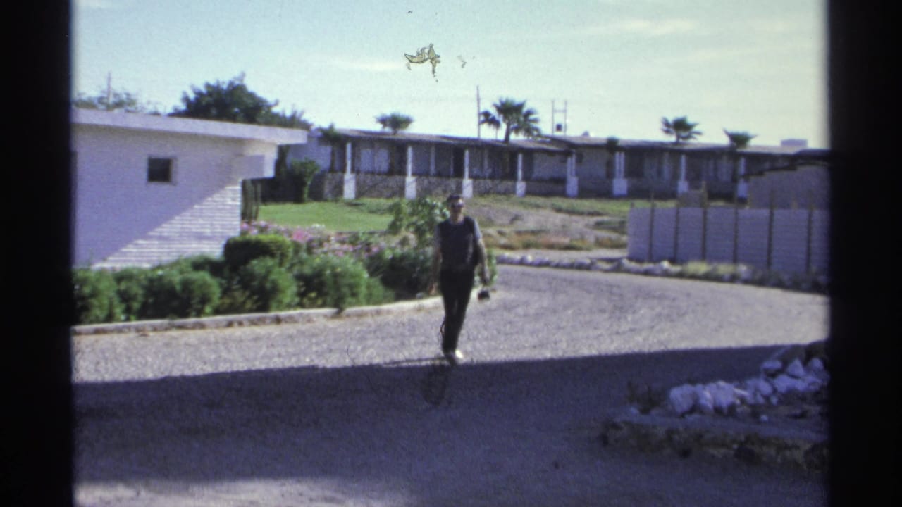 A man approaches on a gravel hotel path in Guaymas, Mexico, with palms, white walls, and resort buildings behind him.
