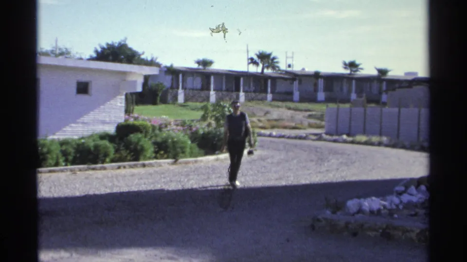 A man approaches on a gravel hotel path in Guaymas, Mexico, with palms, white walls, and resort buildings behind him.