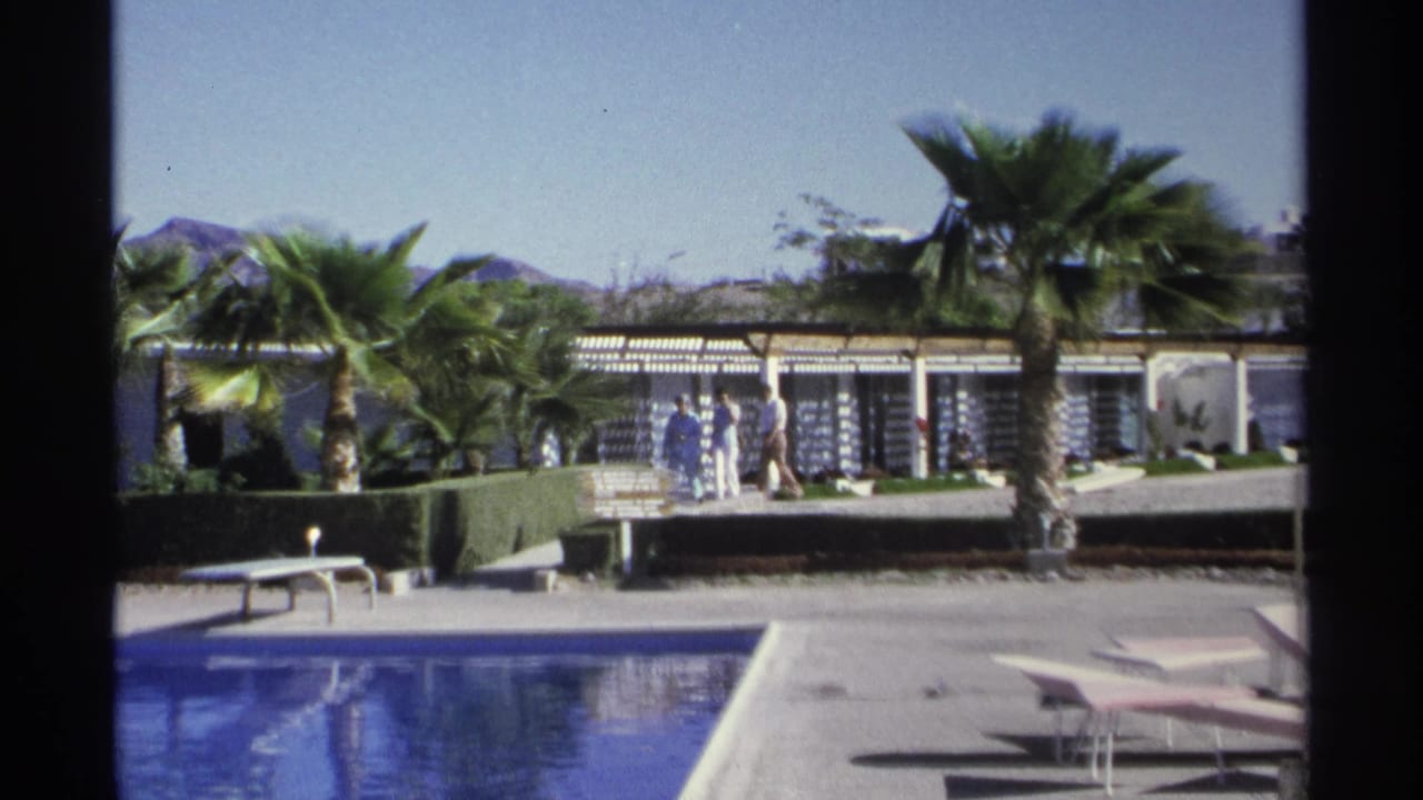 Resort pool with palm trees, hedges, pergola, and small figures walking in the background.