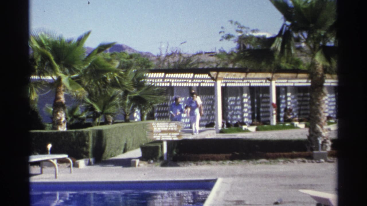 Guaymas, Mexico resort walkway in 1975 with visitors near a pool, palm trees, hedges, and a white building under a pergola.