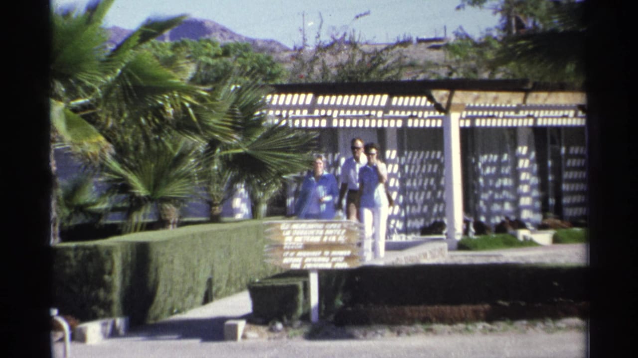 1975 Guaymas resort grounds with a small group walking near a poolside sign, palm trees, hedges, and patterned pergola shade.