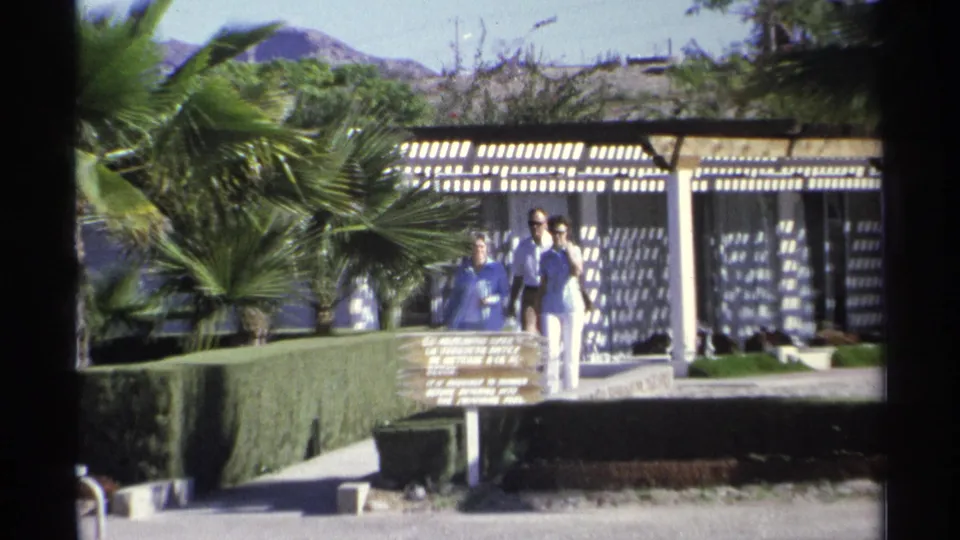 1975 Guaymas resort grounds with a small group walking near a poolside sign, palm trees, hedges, and patterned pergola shade.