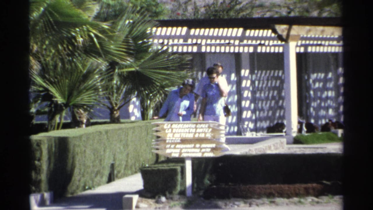 Closer 1975 Guaymas resort view of visitors standing near a poolside sign, framed by palm trees, hedges, and a sunlit pergola.