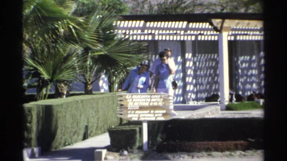 Closer 1975 Guaymas resort view of visitors standing near a poolside sign, framed by palm trees, hedges, and a sunlit pergola.