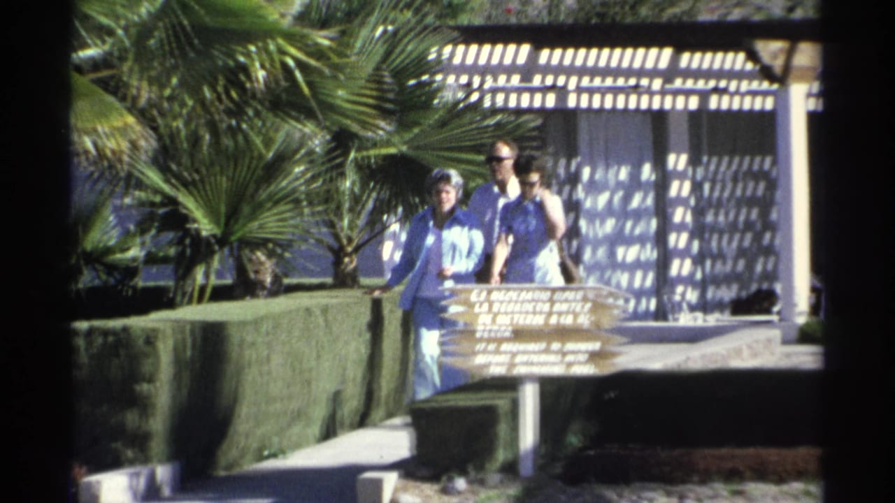 Three adults approach a wooden sign with palms and pergola shade behind them.