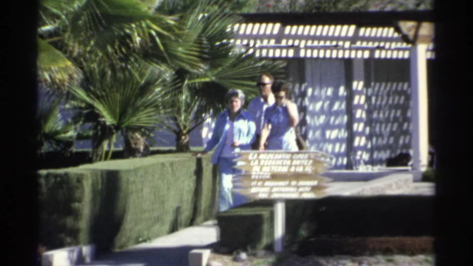 Three adults walk past a wooden resort sign in 1975 Guaymas, with palm fronds, hedges, and pergola shadows in view.