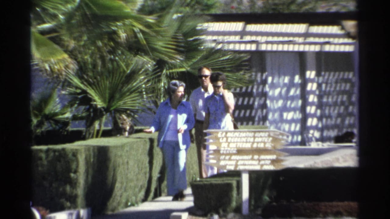 In the Guaymas resort scene, the adults pass close to the wooden sign with hedges, palms, and patterned shade behind them.