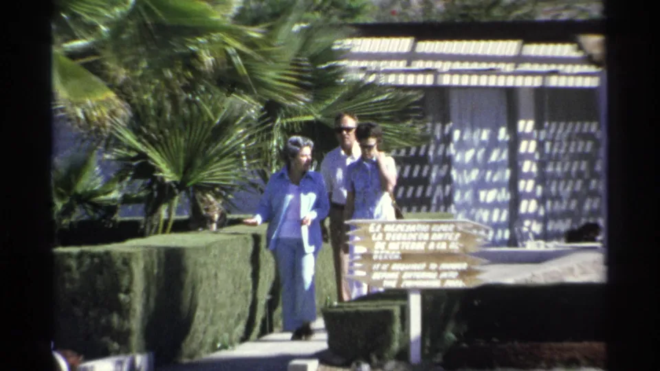 In the Guaymas resort scene, the adults pass close to the wooden sign with hedges, palms, and patterned shade behind them.