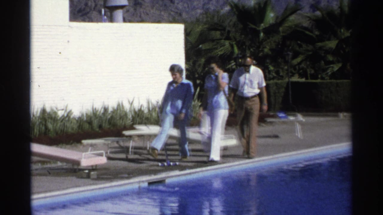 Three adults walk along a Guaymas resort pool in 1975, with a lounge chair, landscaped plants, and a pale wall behind them.