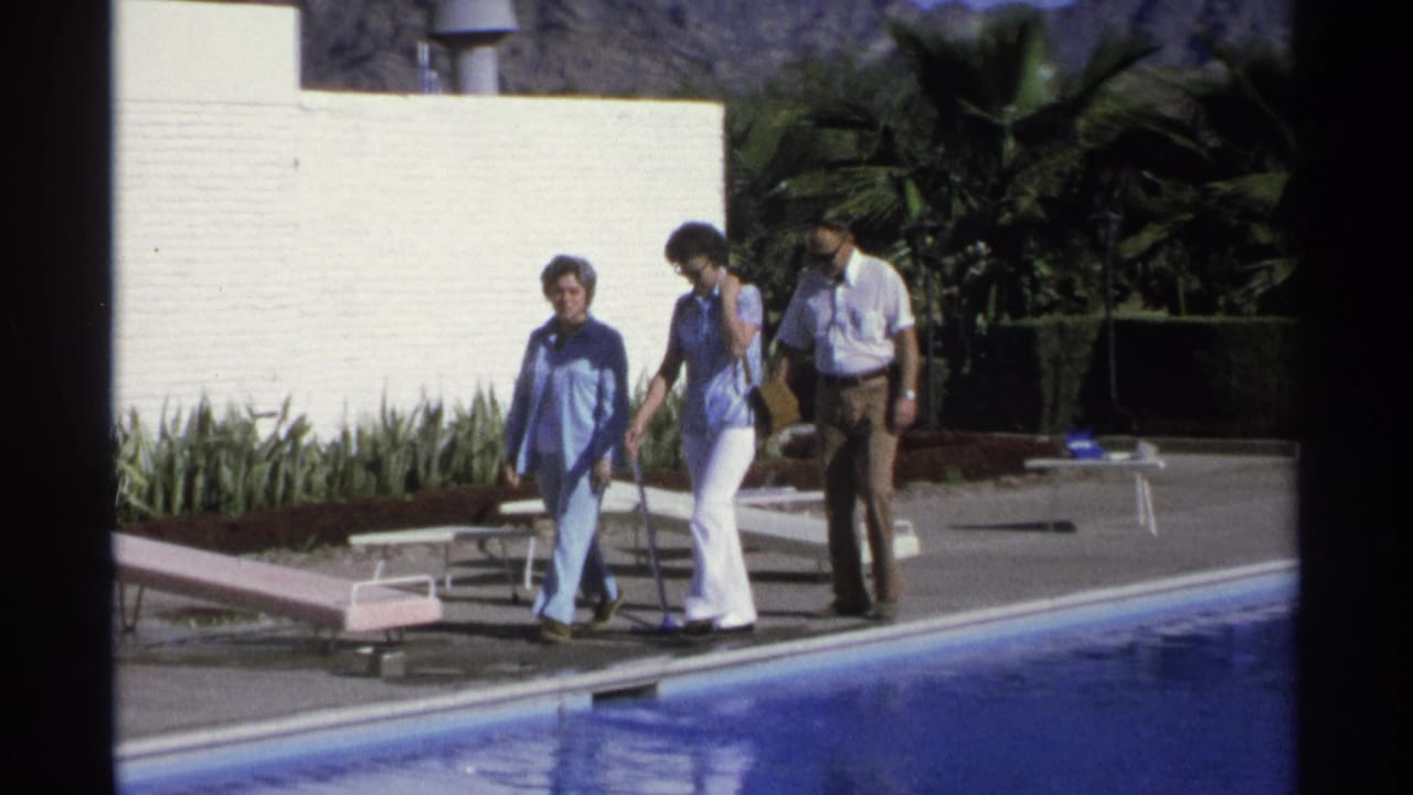 Three adults walking beside a pool with lounge chairs and a white wall behind them.