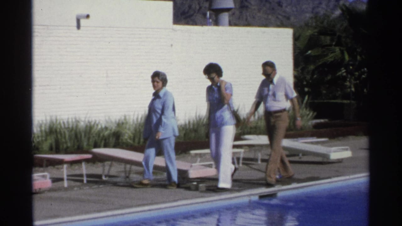 A woman in blue, a woman in white pants, and a man in brown trousers walk past lounge chairs beside a pool in 1975 Guaymas footage.