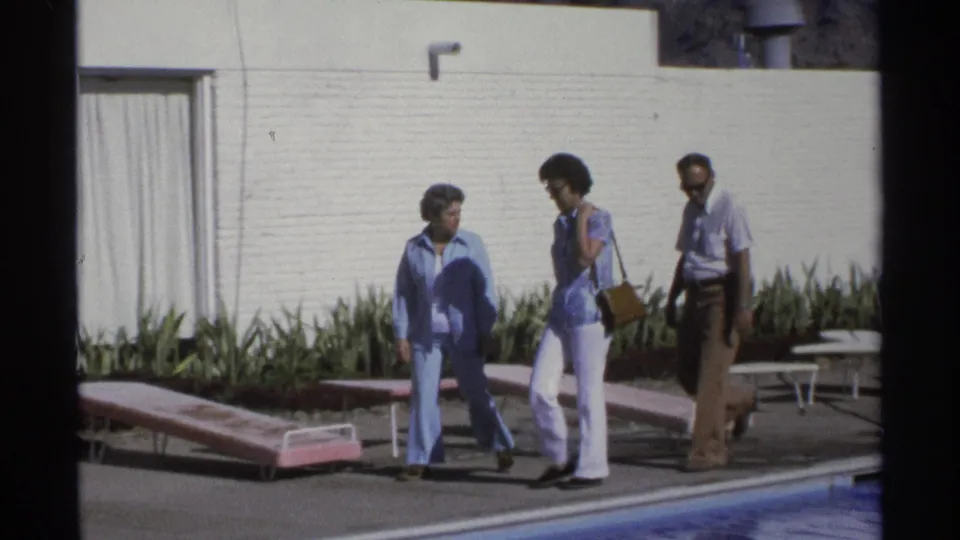 Three adults move along the poolside near pink lounge chairs, a white wall, plants, and a curtained doorway in Guaymas home-movie footage.