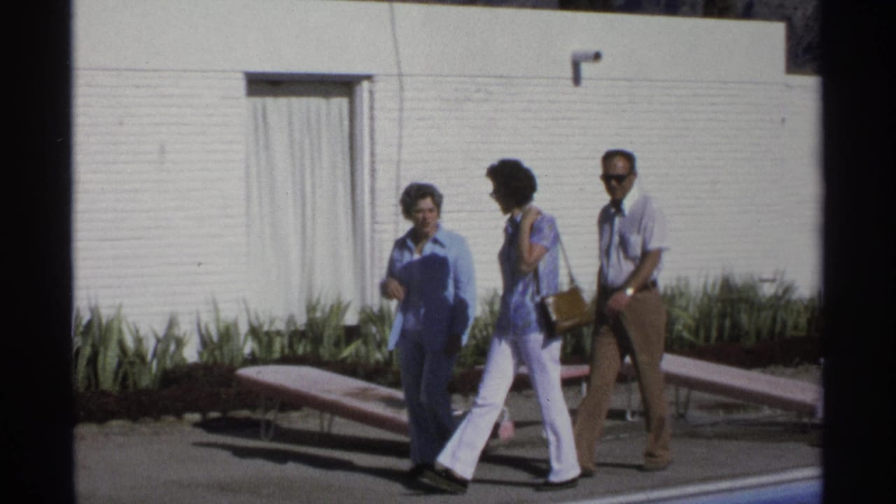 The adults continue past the pool, with the woman in blue leading and lounge chairs and white brick architecture behind them.